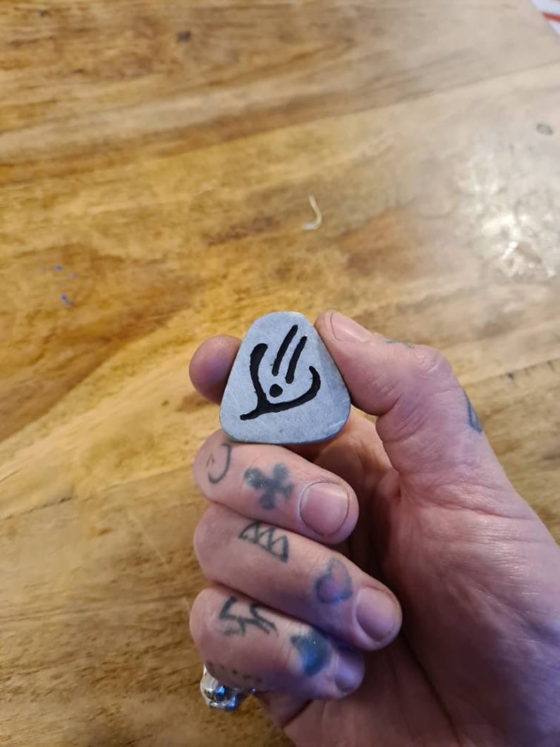 May include: A grey guitar pick with a black design of a stylised hand with three fingers extended. The pick is being held by a hand with tattoos.