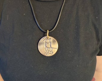 May include: A gold necklace with a round pendant featuring a stylized animal design. The pendant is attached to a black leather cord.