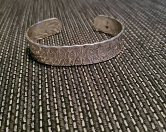 May include: A silver bracelet with a textured, hammered design. The bracelet is open and has a slightly curved shape.