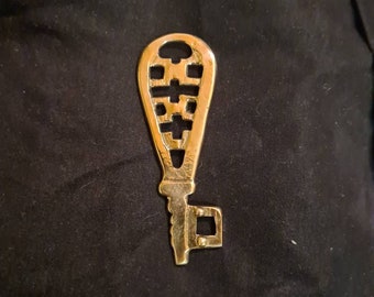 May include: A gold-colored metal key with a decorative, intricate design in the shape of a lacrosse stick. The key has a small, square-shaped hole at the bottom.