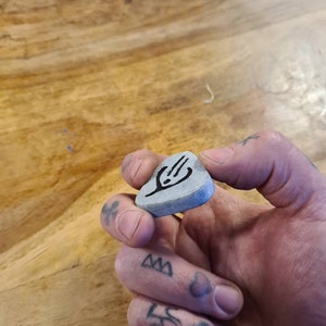 May include: A grey guitar pick with a black design of a hand holding a guitar pick. The pick is being held by a hand with tattoos.