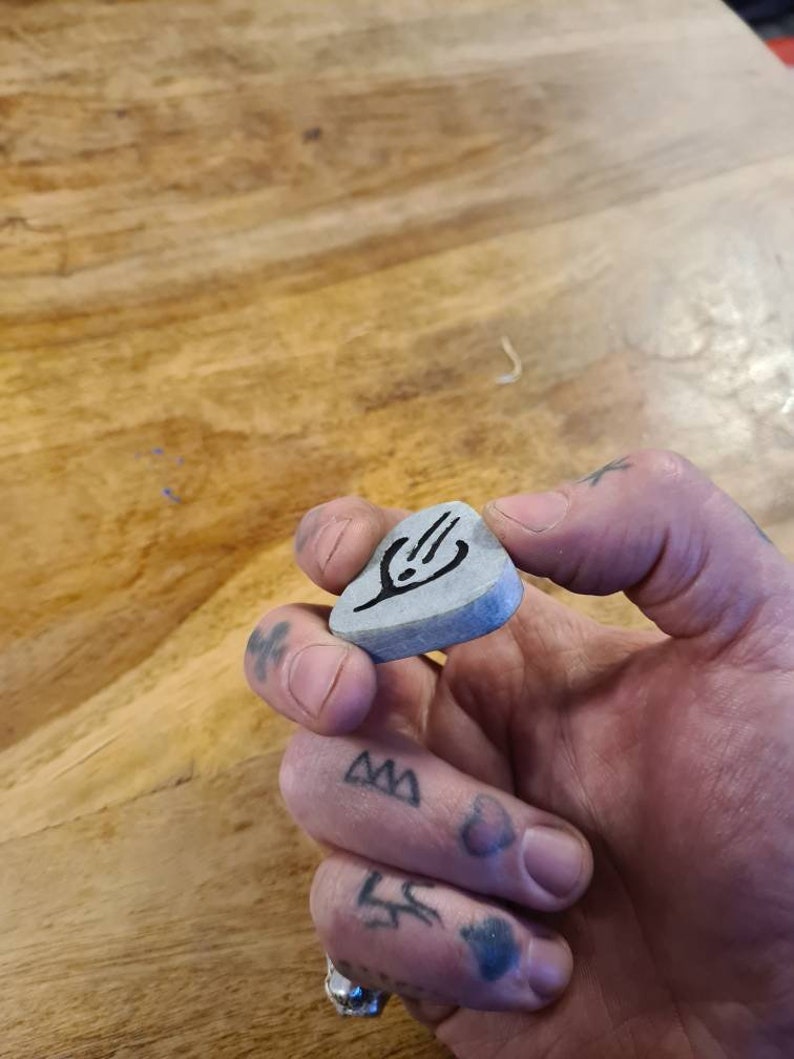 May include: A grey guitar pick with a black design of a hand holding a guitar pick. The pick is being held by a hand with tattoos.