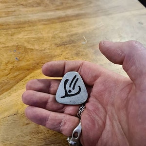 May include: A silver guitar pick with a black design of a stylised hand with three fingers extended.