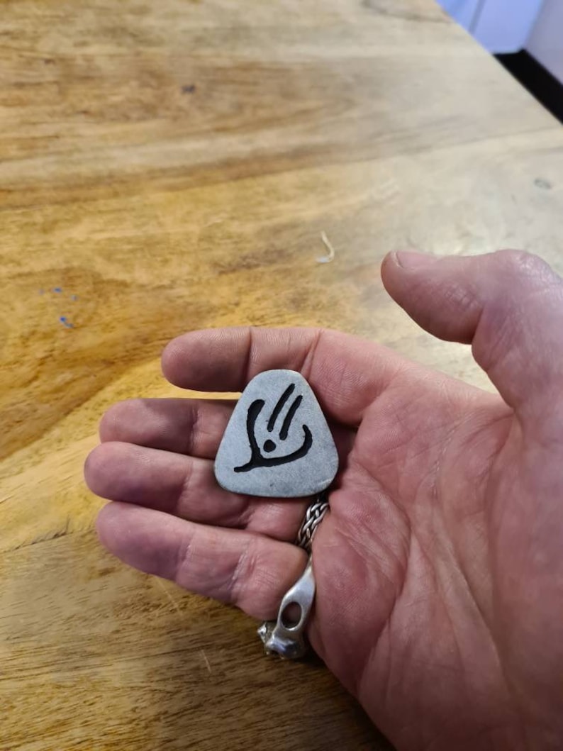 May include: A silver guitar pick with a black design of a stylised hand with three fingers extended.