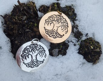 May include: Two silver and gold pendants with a tree of life design. The pendants are on a snowy background.