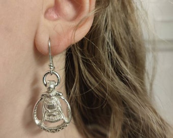 May include: Silver earrings with a detailed design of a stylized figure. The earrings are hanging from a person's ear.