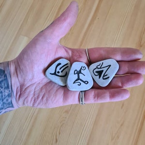 May include: Four grey stones with black markings, resembling runes or symbols, are held in a hand. The stones are smooth and have a natural, unpolished look.
