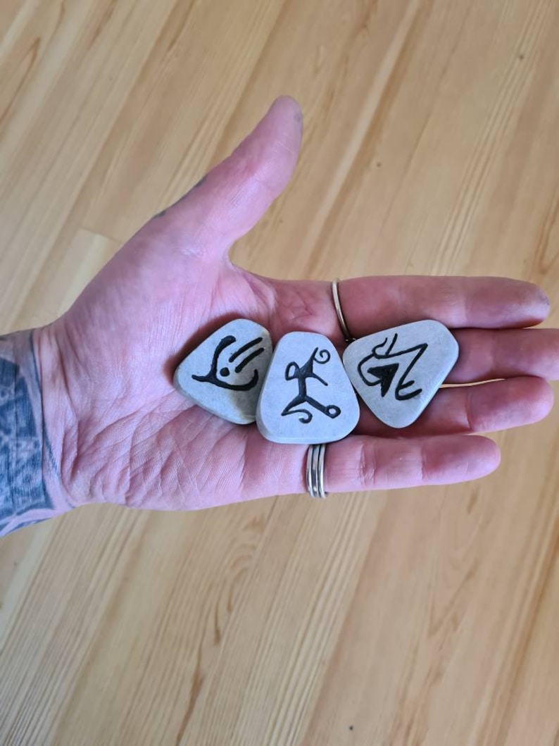 May include: Four grey stones with black markings, resembling runes or symbols, are held in a hand. The stones are smooth and have a natural, unpolished look.