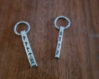 May include: Two silver keychains with a braided ring and a rectangular pendant. One pendant reads "FAITH" in black lettering, and the other has runes. The keychains are displayed on a dark wooden surface.