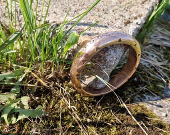 May include: A bronze-colored bangle bracelet with etched geometric designs. The bracelet is resting on a small rock, surrounded by grass and moss. The bracelet's surface reflects sunlight, highlighting its metallic sheen.