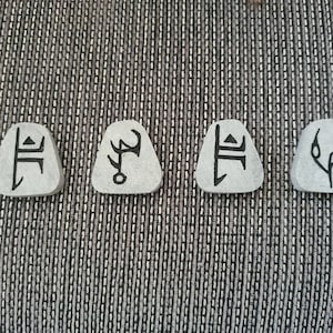 May include: Four small, grey, triangular-shaped stones with black symbols etched on them. The symbols are unique and abstract.