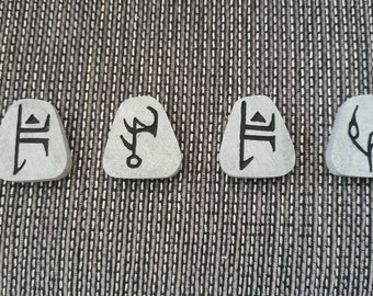 May include: Four small, gray, triangular-shaped stones with black symbols etched on them. The symbols are unique and abstract.