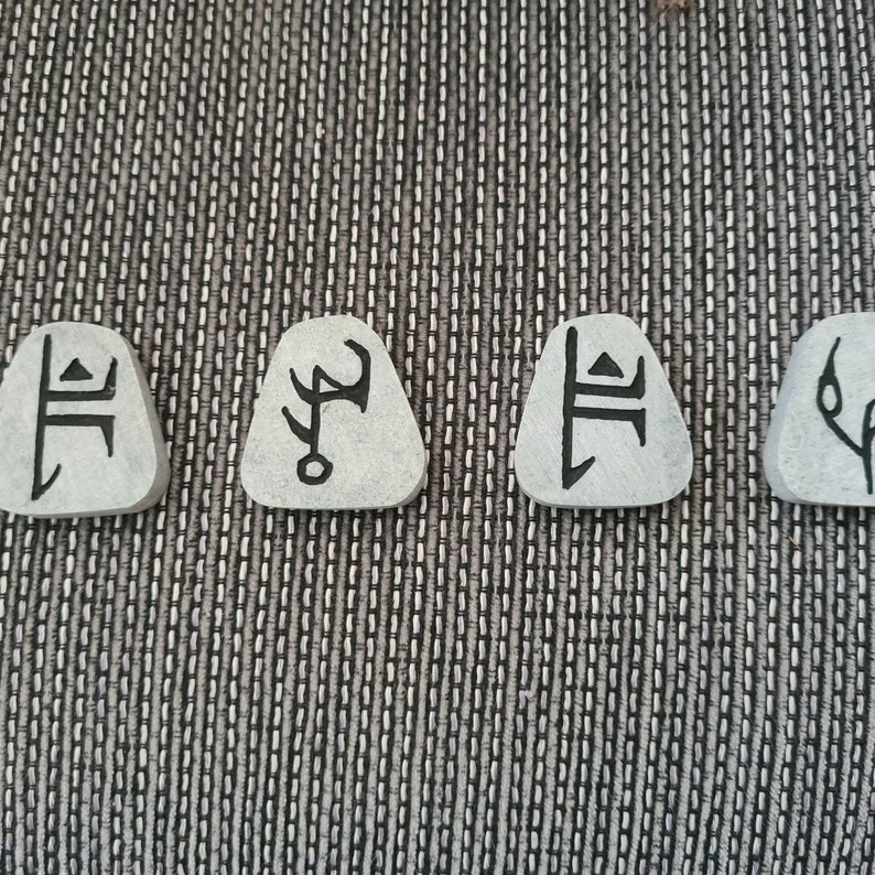 May include: Four small, grey, triangular-shaped stones with black symbols etched on them. The symbols are unique and abstract.
