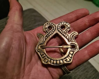 May include: A gold-toned metal belt buckle with a detailed design of two intertwined snakes. The buckle has a circular opening for a belt.