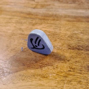 May include: A grey triangular-shaped stone with a black symbol on it. The symbol is a stylised hand with three fingers.