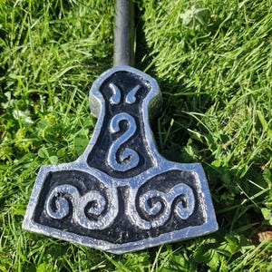 May include: A silver and black Thor's hammer, a replica of the Mjolnir, rests on green grass. The hammer head has detailed spiral and swirl patterns. The handle is a dark grey colour.