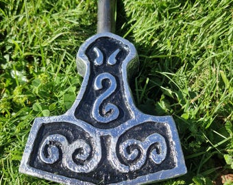 May include: A silver and black Thor's hammer, a replica of the Mjolnir, lies on green grass. The hammer head features intricate spiral and swirl designs. The handle is a dark gray color.