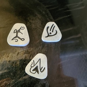 May include: Three grey triangular-shaped tokens with black ink designs. The designs are abstract and resemble symbols.