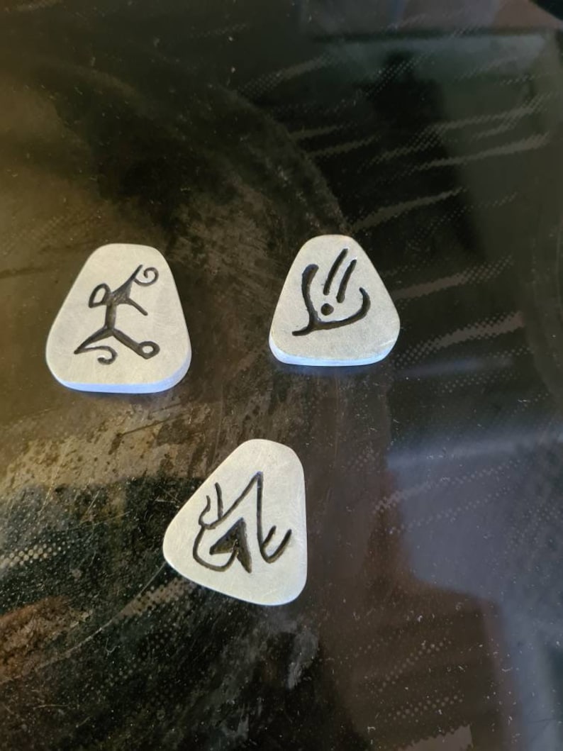 May include: Three grey triangular-shaped tokens with black ink designs. The designs are abstract and resemble symbols.