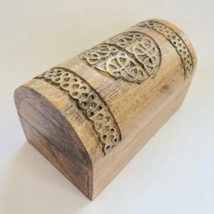 May include: A light brown wooden chest with a curved top and sides. The chest features intricate carved Celtic knot designs around the top and sides. The wood grain is visible, adding to the rustic charm of the chest.