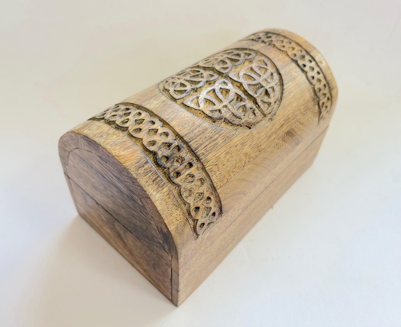 May include: A light brown wooden chest with a curved top and sides. The chest features intricate carved Celtic knot designs around the top and sides. The wood grain is visible, adding to the rustic charm of the chest.
