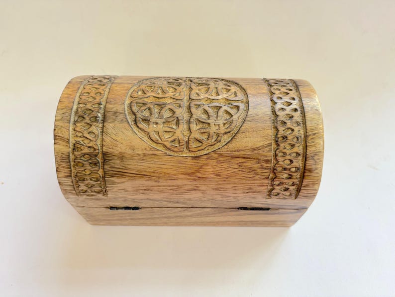 May include: A wooden chest with a carved Celtic knot design. The chest is rectangular with rounded sides and a hinged lid. The wood is light brown in colour. The chest is likely for storing small items.