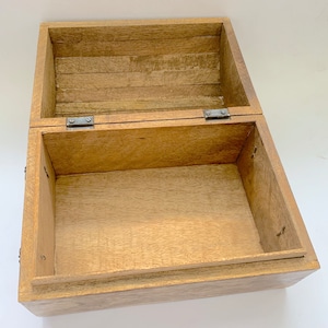 May include: An open, rectangular wooden box with a hinged lid. The box is made of light-coloured wood with visible grain and texture. Metal hinges and small nails secure the lid and box structure. The interior is empty, revealing the box's construction.