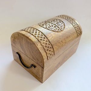 May include: A wooden chest with a curved top and detailed Celtic knot carvings. The chest is made of light-coloured wood and has a black handle. The carved details are highlighted with a lighter colour.