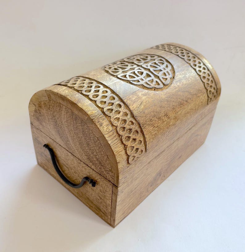 May include: A wooden chest with a curved top and detailed Celtic knot carvings. The chest is made of light-coloured wood and has a black handle. The carved details are highlighted with a lighter colour.