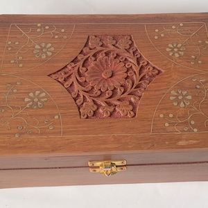 May include: A rectangular wooden box with a hinged lid. The box is a warm brown colour and features intricate floral carvings and brass accents. The centre of the lid has a carved floral design, and the corners have floral patterns.