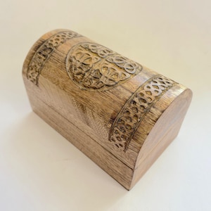 May include: A light brown wooden chest with a curved top, featuring carved Celtic knot patterns. The rectangular base supports the chest. The carved details are darker than the wood. This chest is likely used for storing small items, such as jewelry.
