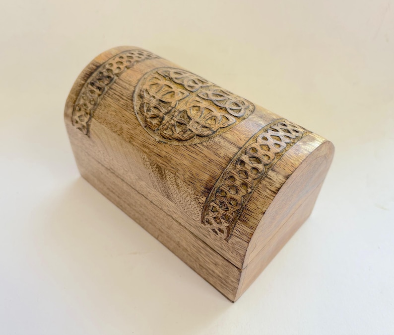 May include: A light brown wooden chest with a curved top, featuring carved Celtic knot patterns. The rectangular base supports the chest. The carved details are darker than the wood. This chest is likely used for storing small items, such as jewelry.