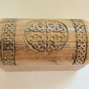 May include: A wooden chest with a carved Celtic knot design on the front and decorative borders. The chest is a light brown colour and has a curved top. The wood grain is visible.