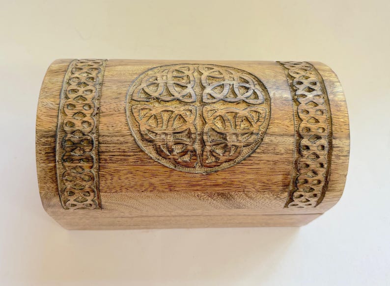 May include: A wooden chest with a carved Celtic knot design on the front and decorative borders. The chest is a light brown colour and has a curved top. The wood grain is visible.