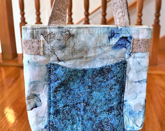 May include: A quilted tote bag with a front pocket. The bag features a blue and white abstract pattern on the main body and a darker blue, swirling pattern on the pocket. The handles and trim are a textured, neutral colour.