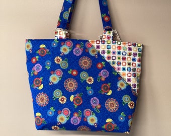 Könnte beinhalten: Eine blaue Tragetasche mit einem Blumenmuster auf der einen Seite und einem geometrischen Muster auf der anderen Seite. Die Tasche hat passende blaue Griffe und ist aus Stoff. Das Blumenmuster enthält bunte Blumen.