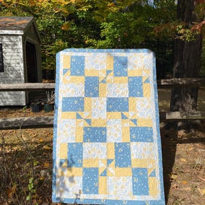 May include: A handmade quilt with a patchwork design. The quilt features a pattern of alternating blue and yellow squares, with white squares adorned with stars and crescent moons. The quilt is displayed outdoors, leaning against a wooden fence.