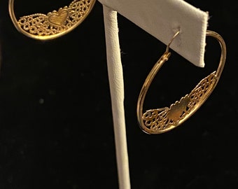 May include: A pair of gold hoop earrings with a delicate heart design. The earrings are displayed on a white stand against a black background.