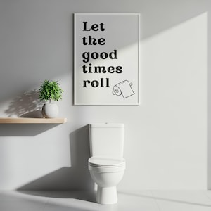 Könnte beinhalten: Ein weiß gerahmter Druck mit dem Text "Let the good times roll" und einer Toilettenpapierrollen-Illustration hängt über einer weißen Toilette. Eine kleine Topfpflanze steht links auf einem Holzregal. Das Badezimmer ist weiß gestrichen.