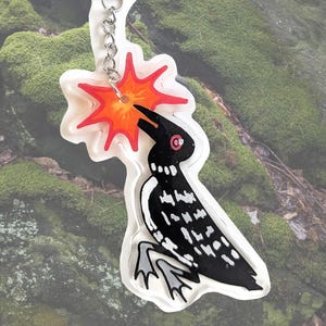 Puede incluir: Un llavero de acrílico transparente con un pájaro estilizado en blanco y negro con un ojo rojo y un diseño de explosión rojo y naranja. El llavero tiene una cadena y un cierre plateados para sujetarlo a llaves o bolsos.