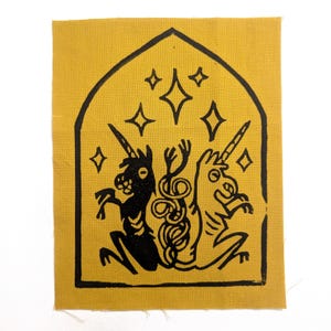 May include: Mustard yellow fabric patch with a black illustration of two unicorns facing each other. The unicorns are inside a black arched frame with star and diamond shapes above them. The patch has a raw edge.