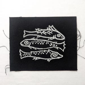 May include: A black fabric patch featuring a white design of three stylised fish swimming in a row. The fish are outlined in white and have a simple, graphic design.