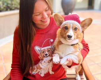 May include: A red long-sleeve sweater featuring a graphic of a Corgi wearing a Santa hat, paired with a small Corgi dog wearing a matching red and white crocheted Santa hat. The sweater has a crew neckline and a knit texture throughout.