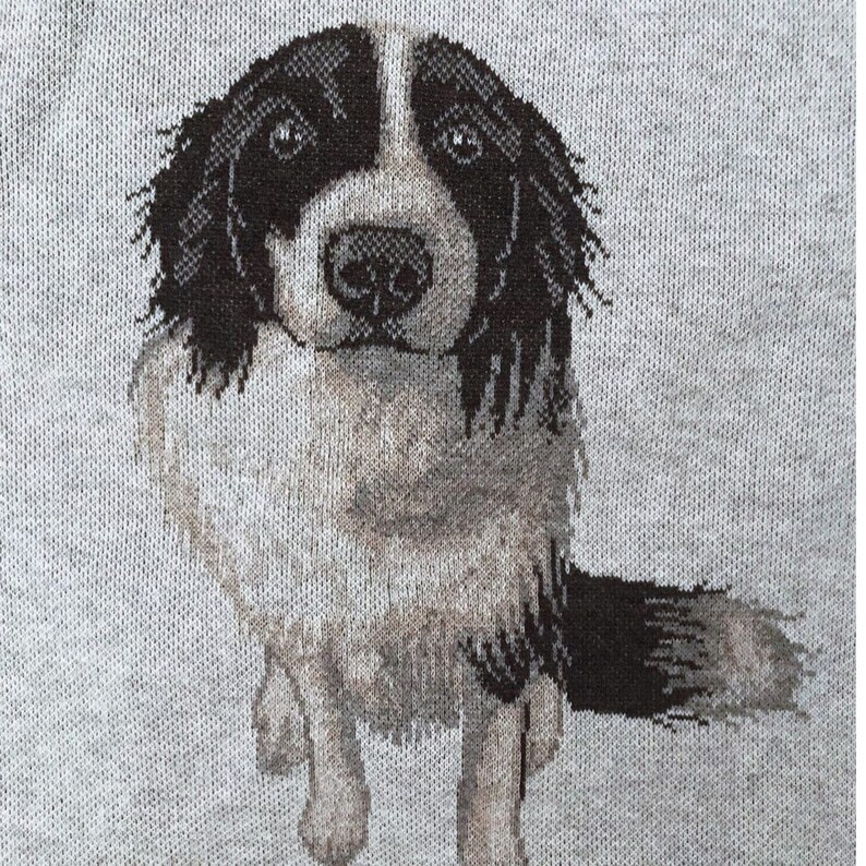 May include: A light gray knit fabric featuring a detailed jacquard-woven graphic of a dog with black and white fur. The dog has a white blaze on its face, dark ears, and a fluffy tail, depicted in a seated position facing forward.
