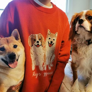 May include: A bright orange long-sleeve crewneck sweater featuring a knitted graphic of two dogs sitting side-by-side. Below the dogs, the text Alyx Moco is embroidered in white. The sweater has a ribbed collar and cuffs.