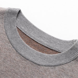 May include: A crew neck sweater featuring a textured knit fabric in a muted brown tone. The garment has a ribbed collar in a contrasting light gray color, showcasing a detailed view of the neckline and the woven material construction.