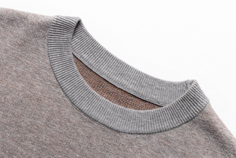 May include: A crew neck sweater featuring a textured knit fabric in a muted brown tone. The garment has a ribbed collar in a contrasting light gray color, showcasing a detailed view of the neckline and the woven material construction.