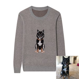 May include: A long-sleeved crew neck sweater in a heathered taupe color. The center of the chest features a knitted graphic of a black and white dog with tan markings, wearing a brown collar, matching the dog shown in the inset photograph.