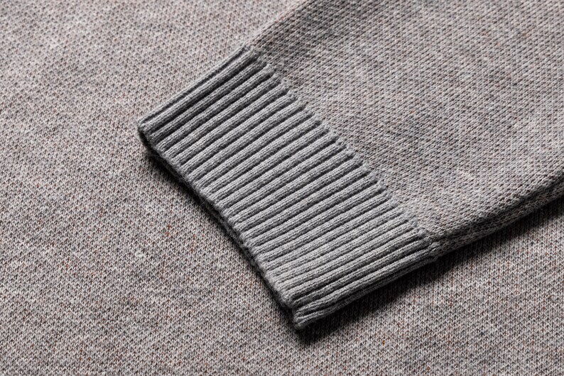 May include: A gray knit sweater sleeve featuring a textured, heathered fabric pattern and a ribbed cuff. The material displays a fine, consistent weave with subtle flecks of darker color throughout the gray base, ending in a structured, elasticized ribbed knit cuff.