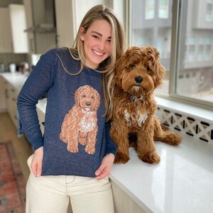 May include: A dark blue long-sleeve sweater featuring a graphic illustration of a brown and white curly-haired dog sitting in the center. The sweater has a crew neckline and a knit texture, showcasing a detailed depiction of a dog breed.